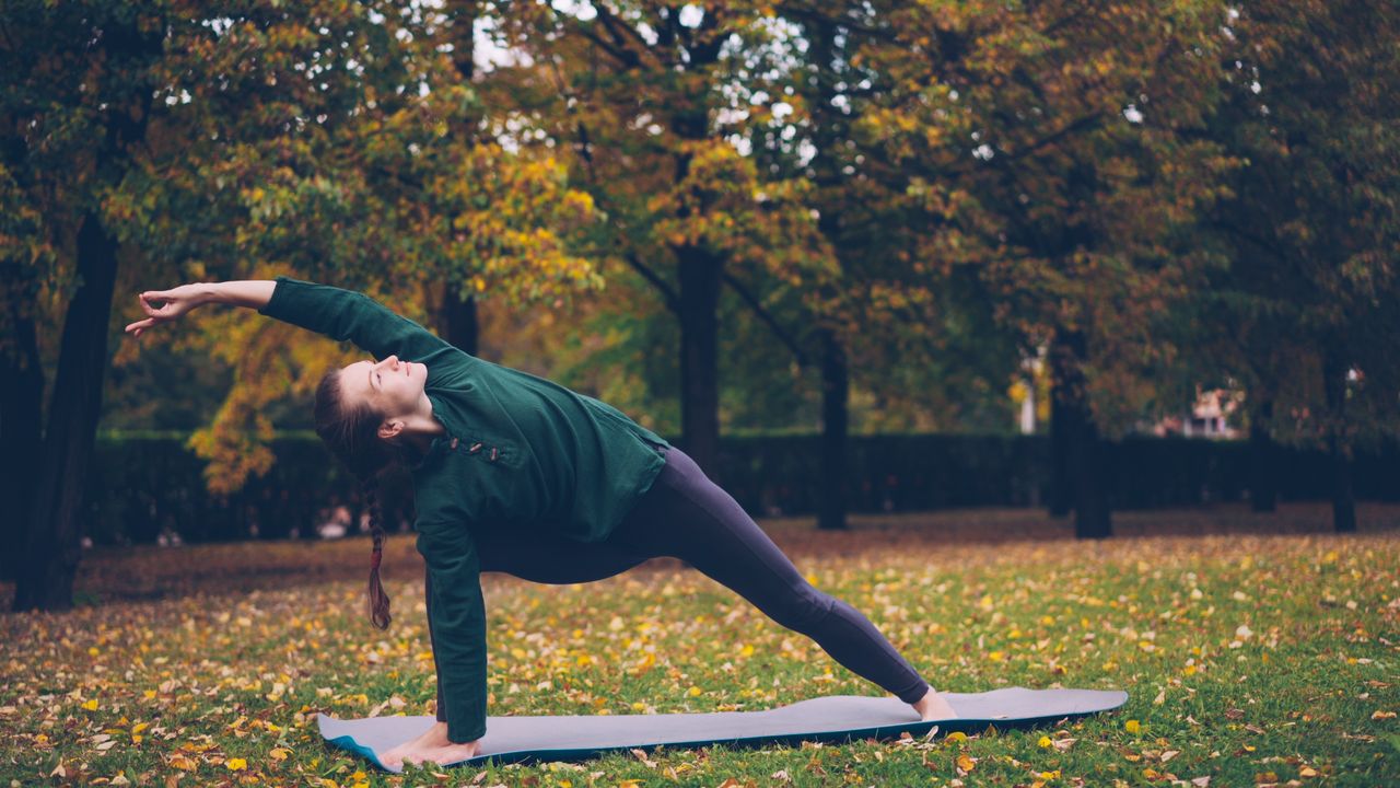 Woman doing a yoga pose on a mat in a park surrounded by autumn trees.